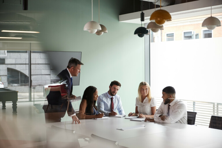 group meeting around table at graduate recruitment assessment day and collaborating on task whilst being observed by mature businessman